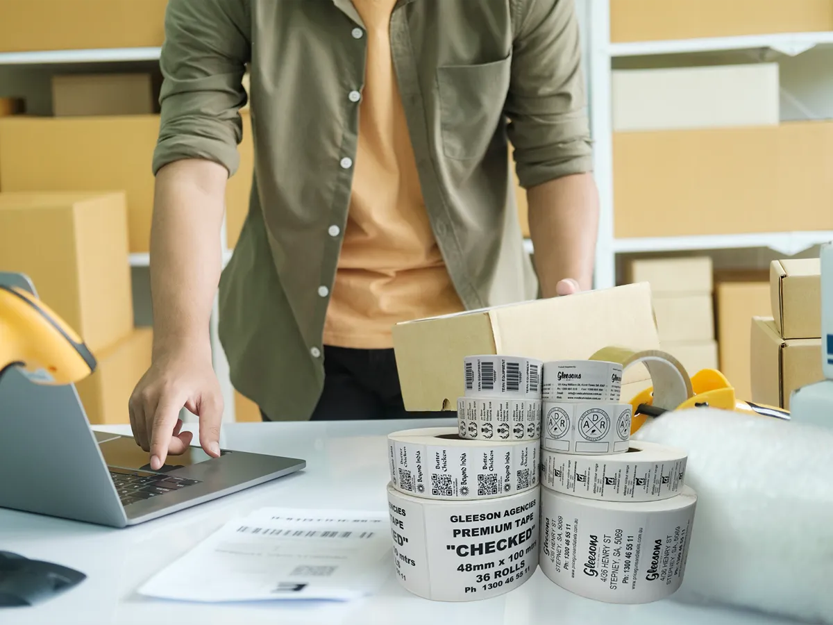 Person using a laptop with rolls of labels on a table, surrounded by boxes.